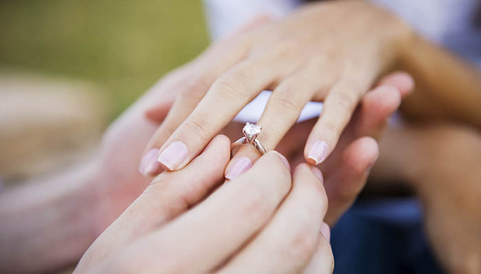 Young man is seen slipping an engagement ring on his girlfriend's ring finger.